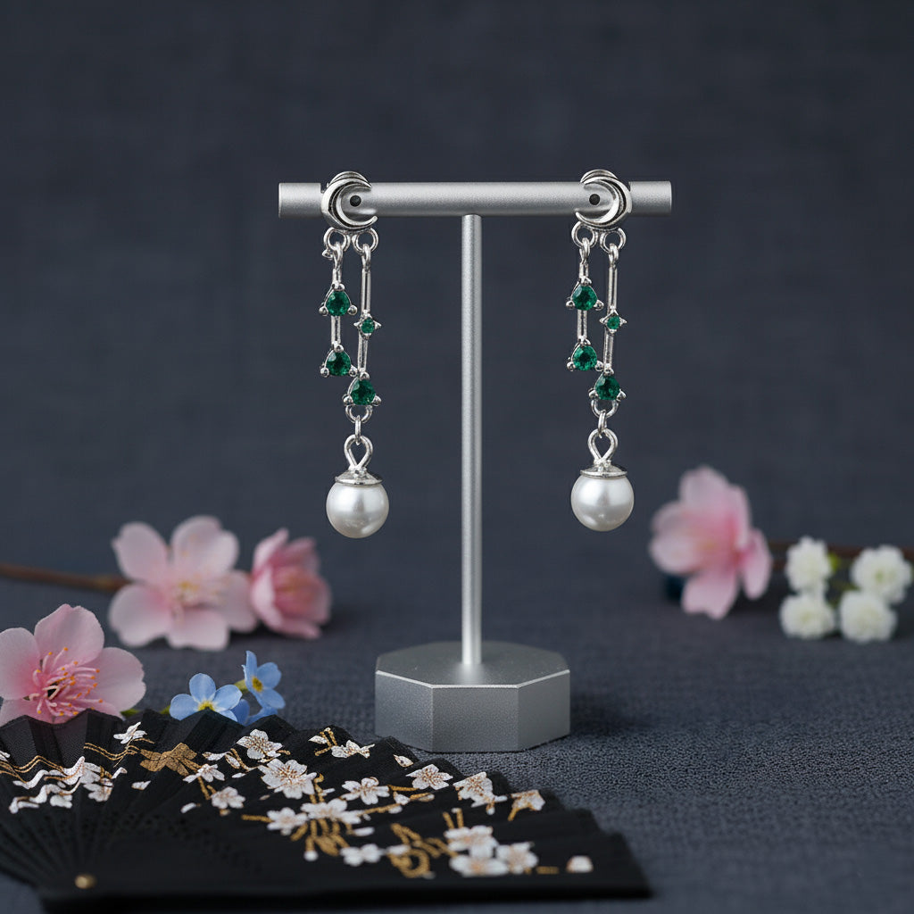 Pair of silver earrings with green gemstones and pearl-like beads on a stand with flowers in the background.