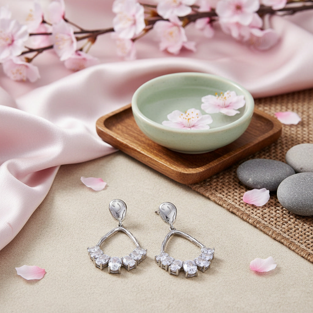 Silver earrings with clear stones on a soft surface with cherry blossoms and a small bowl.