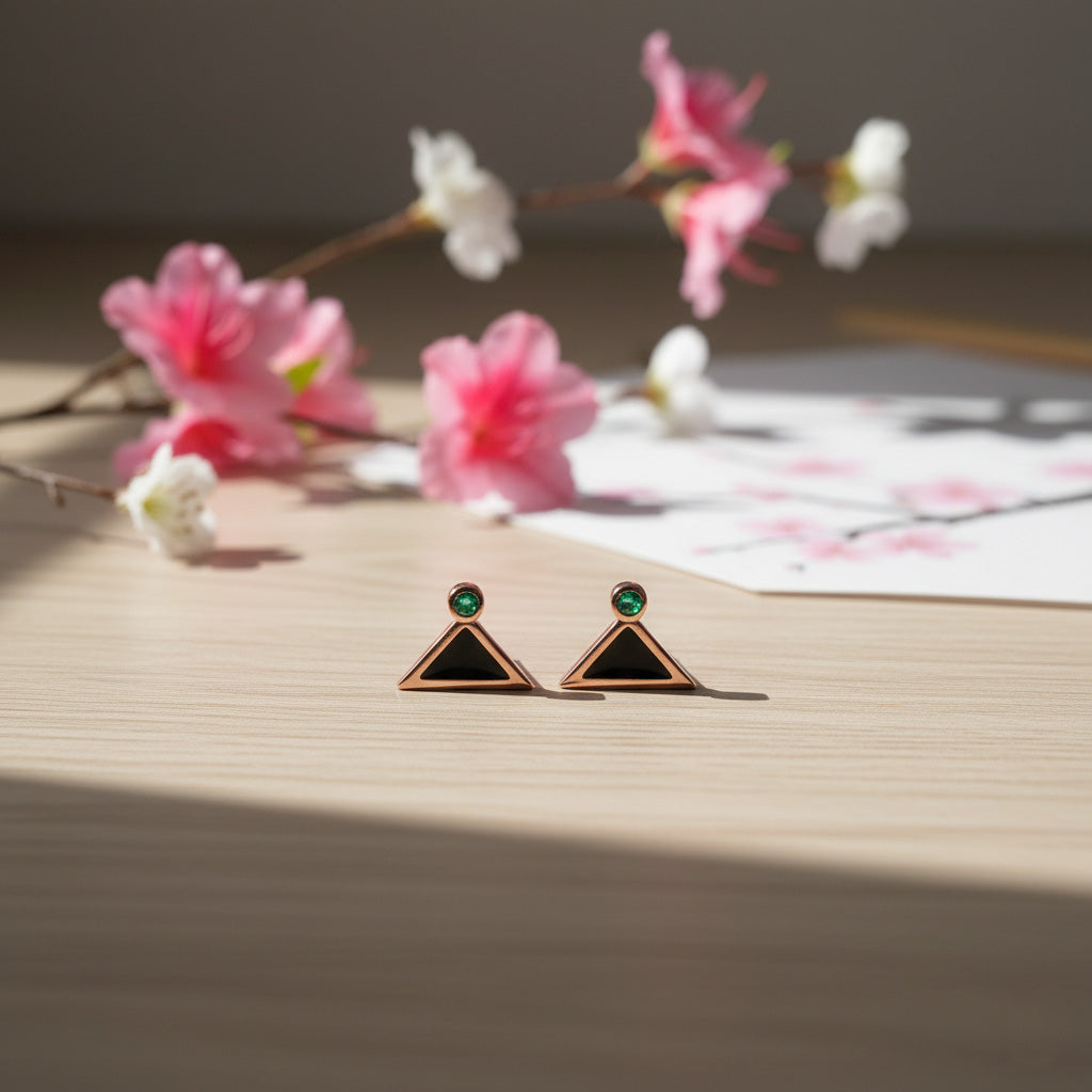 Pair of triangle-shaped earrings with green gemstones on a wooden surface with flowers in the background.