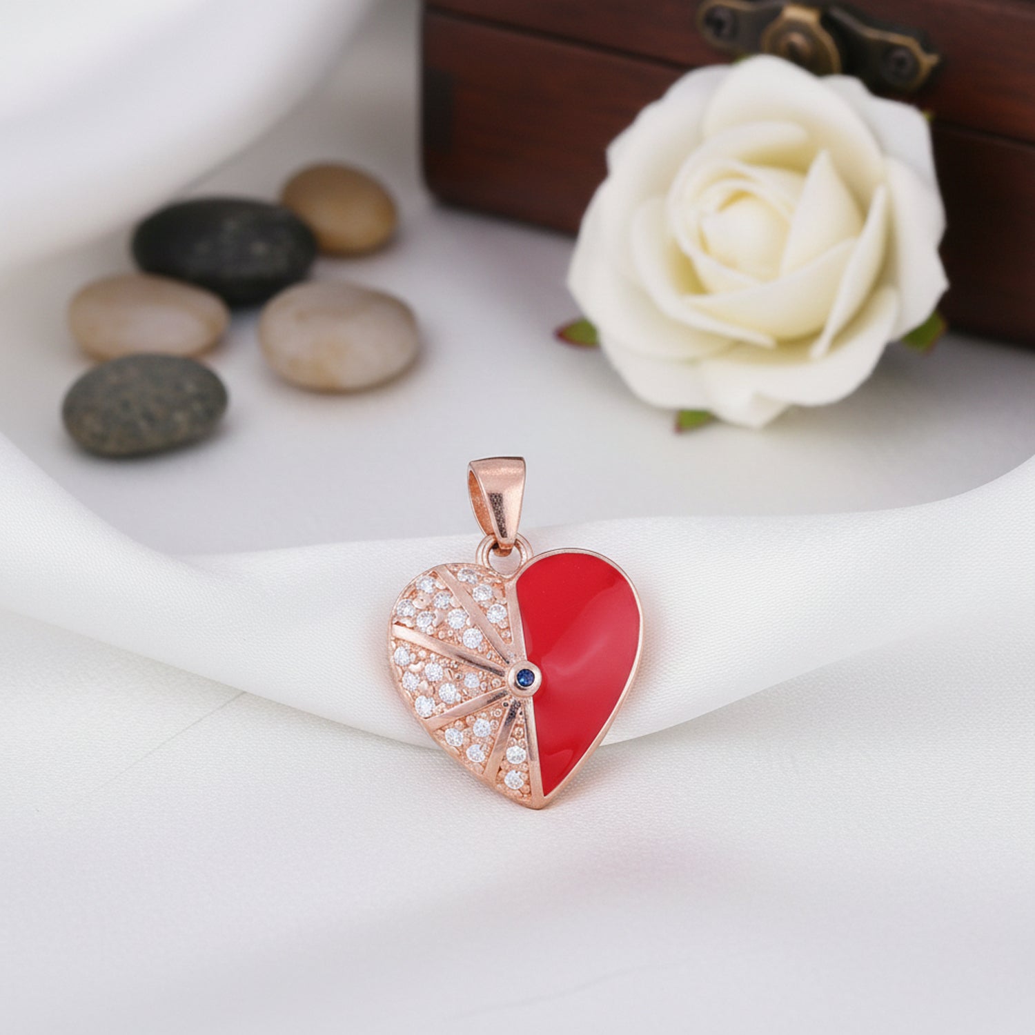 Heart-shaped pendant with red center and rose gold border on a white surface with stones and a rose.