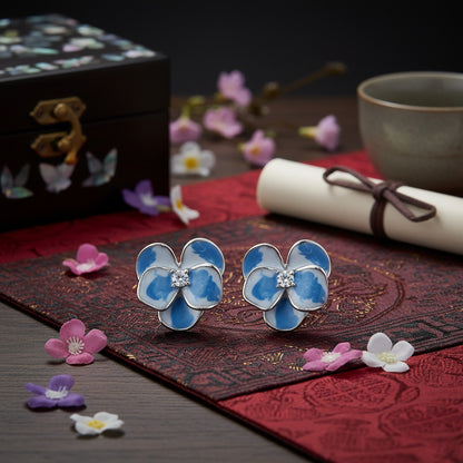 Blue flower-shaped earrings on a decorative mat with flowers and a tea cup in the background
