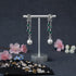 Pair of silver earrings with green gemstones and pearl-like beads on a stand with flowers in the background.