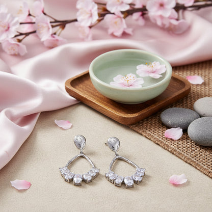 Silver earrings with clear stones on a soft surface with cherry blossoms and a small bowl.