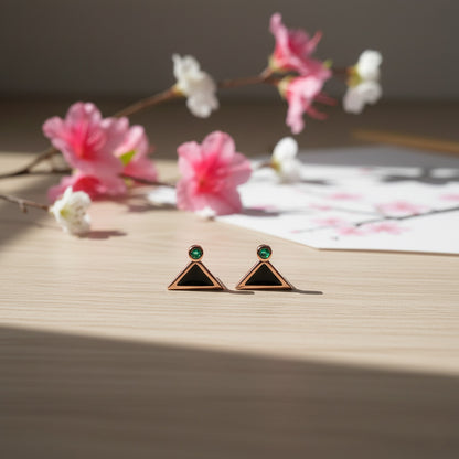 Pair of triangle-shaped earrings with green gemstones on a wooden surface with flowers in the background.