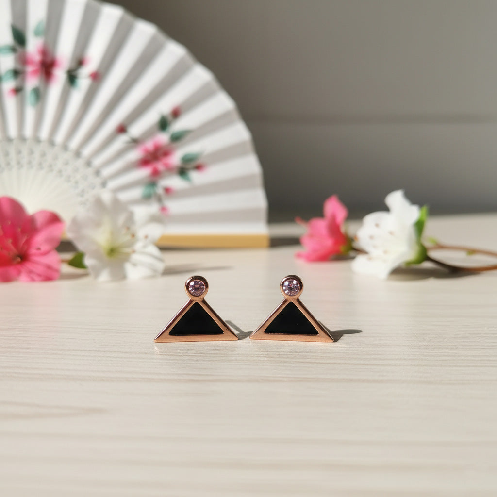 Rose gold triangle-shaped earrings with black stones on a light surface with flowers in the background