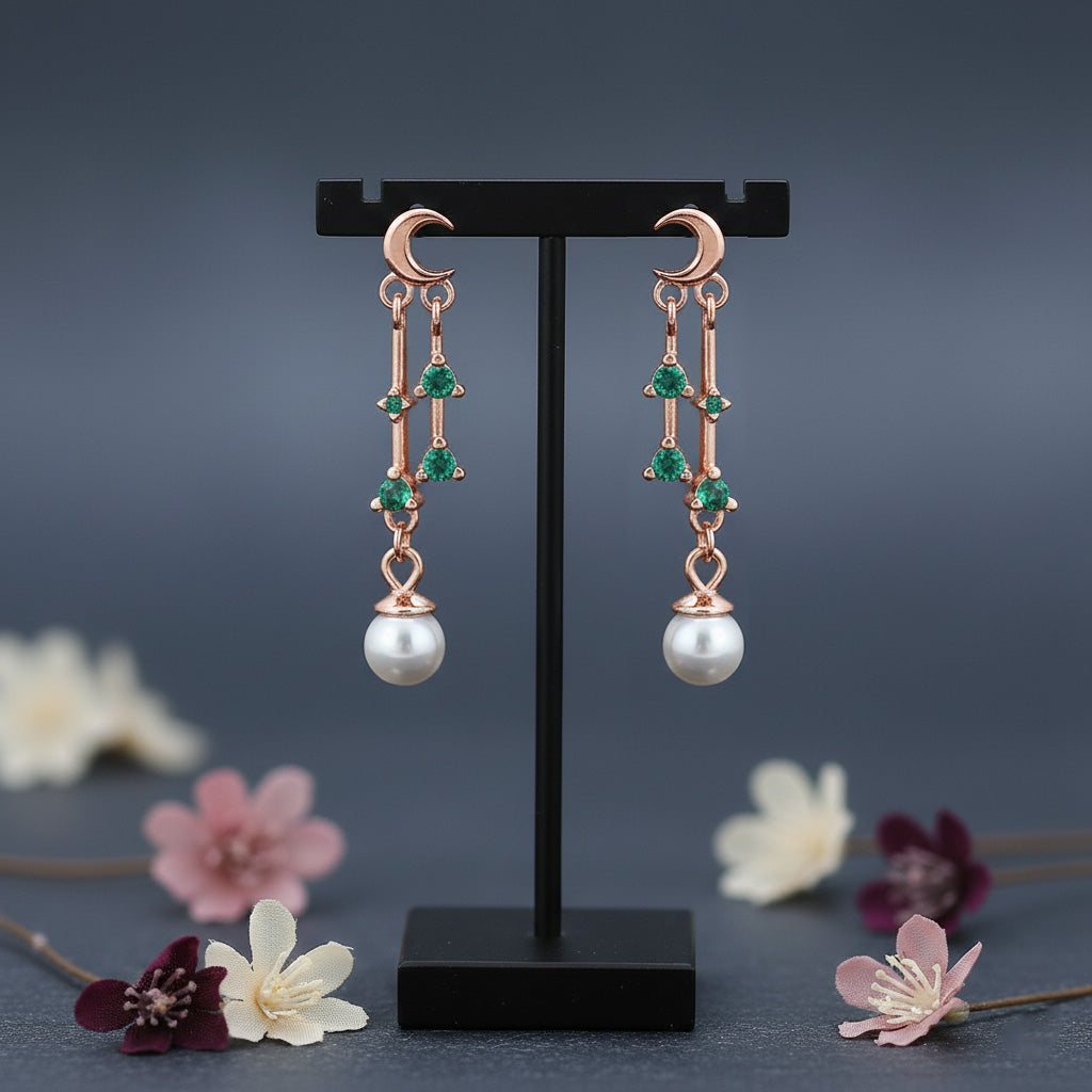 Rose gold earrings with green gemstones and pearl drops on a black stand against a dark background with flowers.