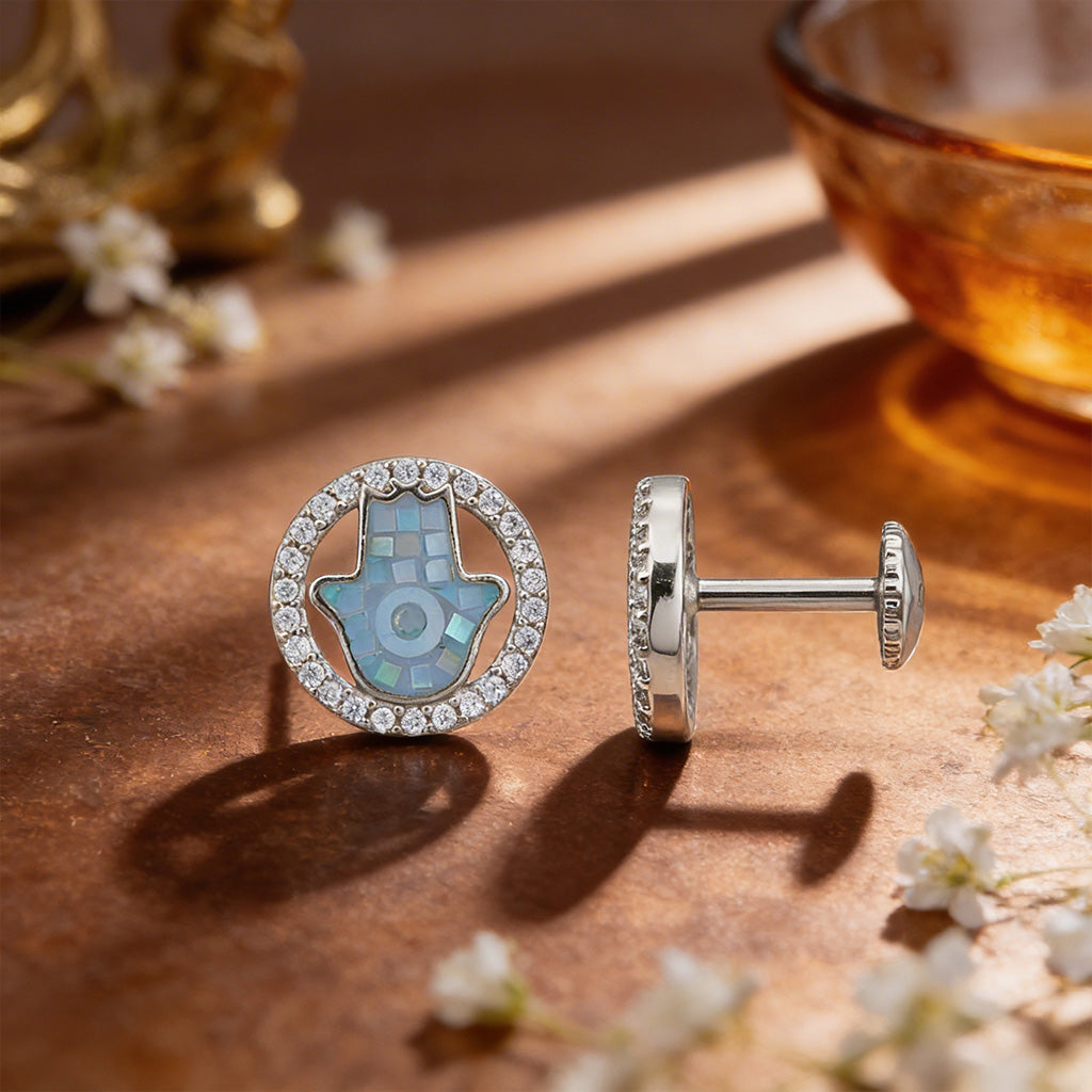 Silver earrings with a Hamsa hand design on a wooden surface with flowers and a bowl.
