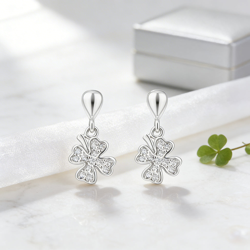 Silver four-leaf clover earrings on a white surface with a green leaf for scale.