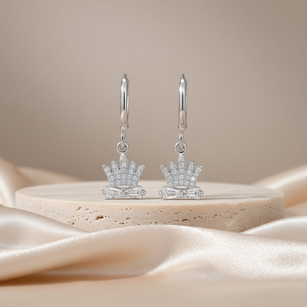 Silver crown-shaped earrings on a beige stone surface with a soft beige background