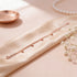 Rose gold bracelet on a beige fabric surface with a pearl necklace and small dish in the background.