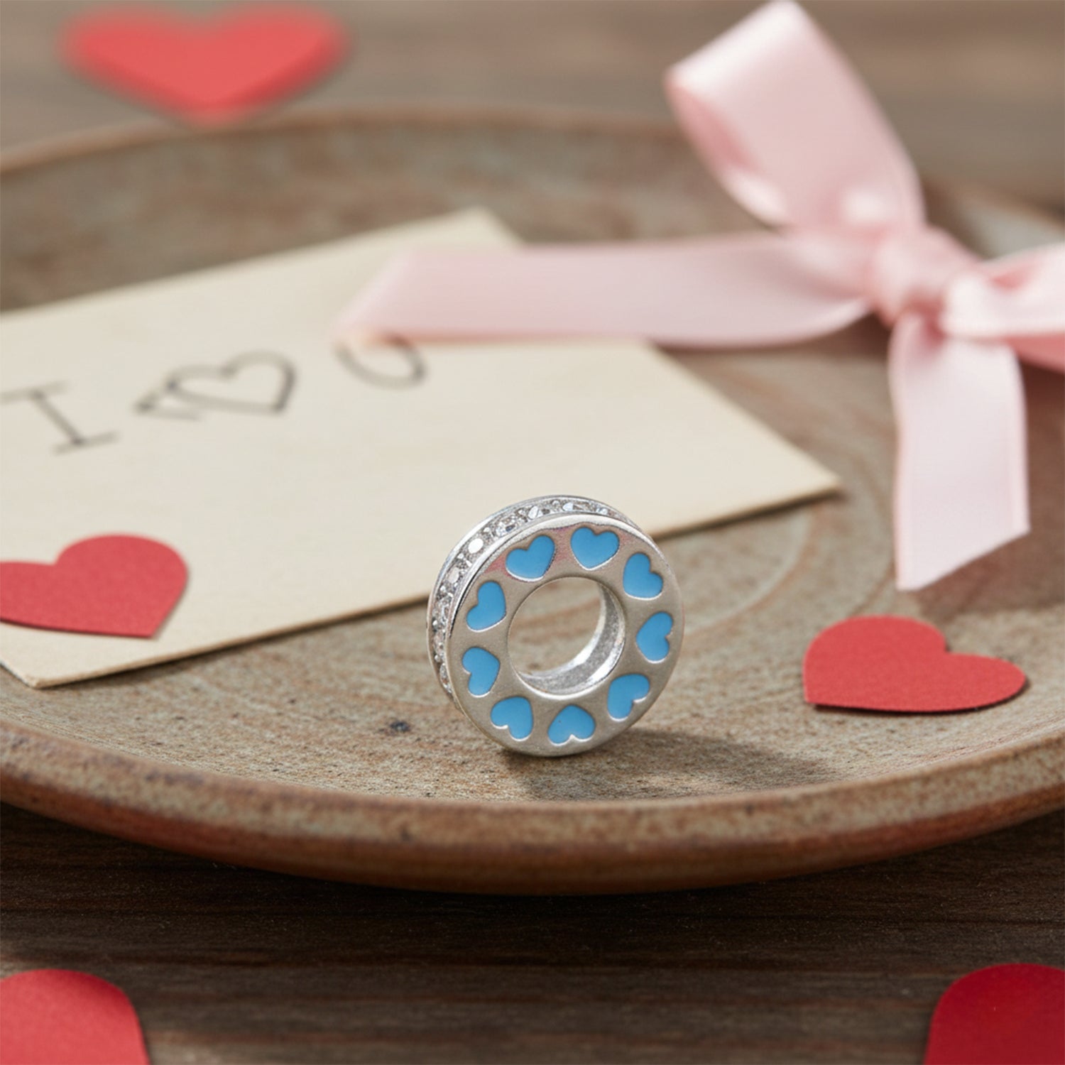 Silver charm with blue hearts on a wooden surface with red hearts and a pink ribbon.