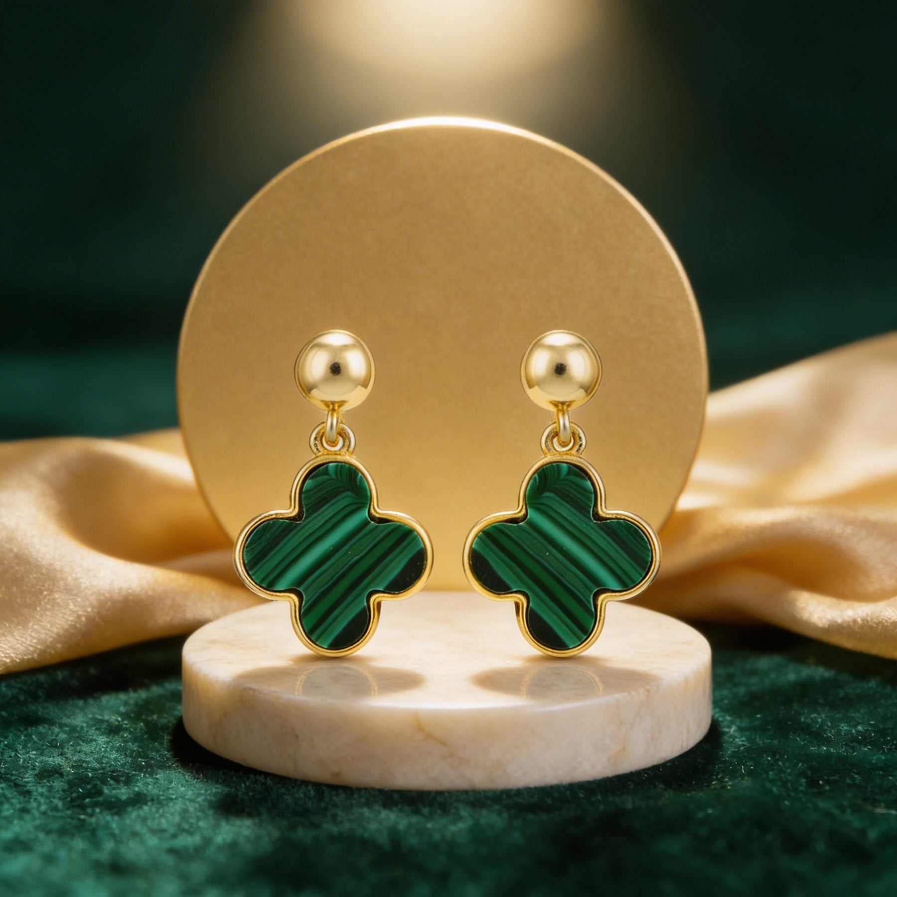 Gold earrings with green clover-shaped stones on a marble stand with a gold and green fabric background.