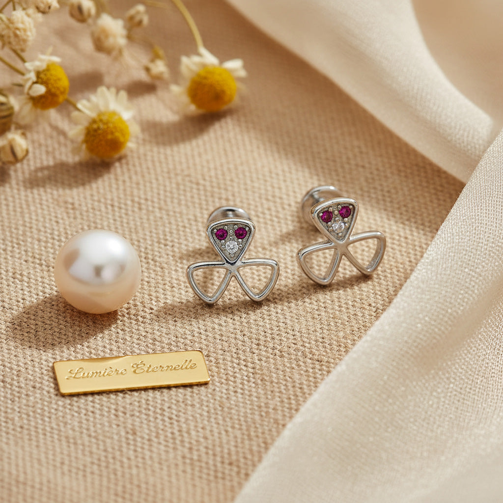 Silver bow-shaped earrings with red gemstones on a beige fabric background with a pearl and dried flowers.