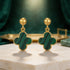 Gold earrings with green clover-shaped drops on a wooden surface.